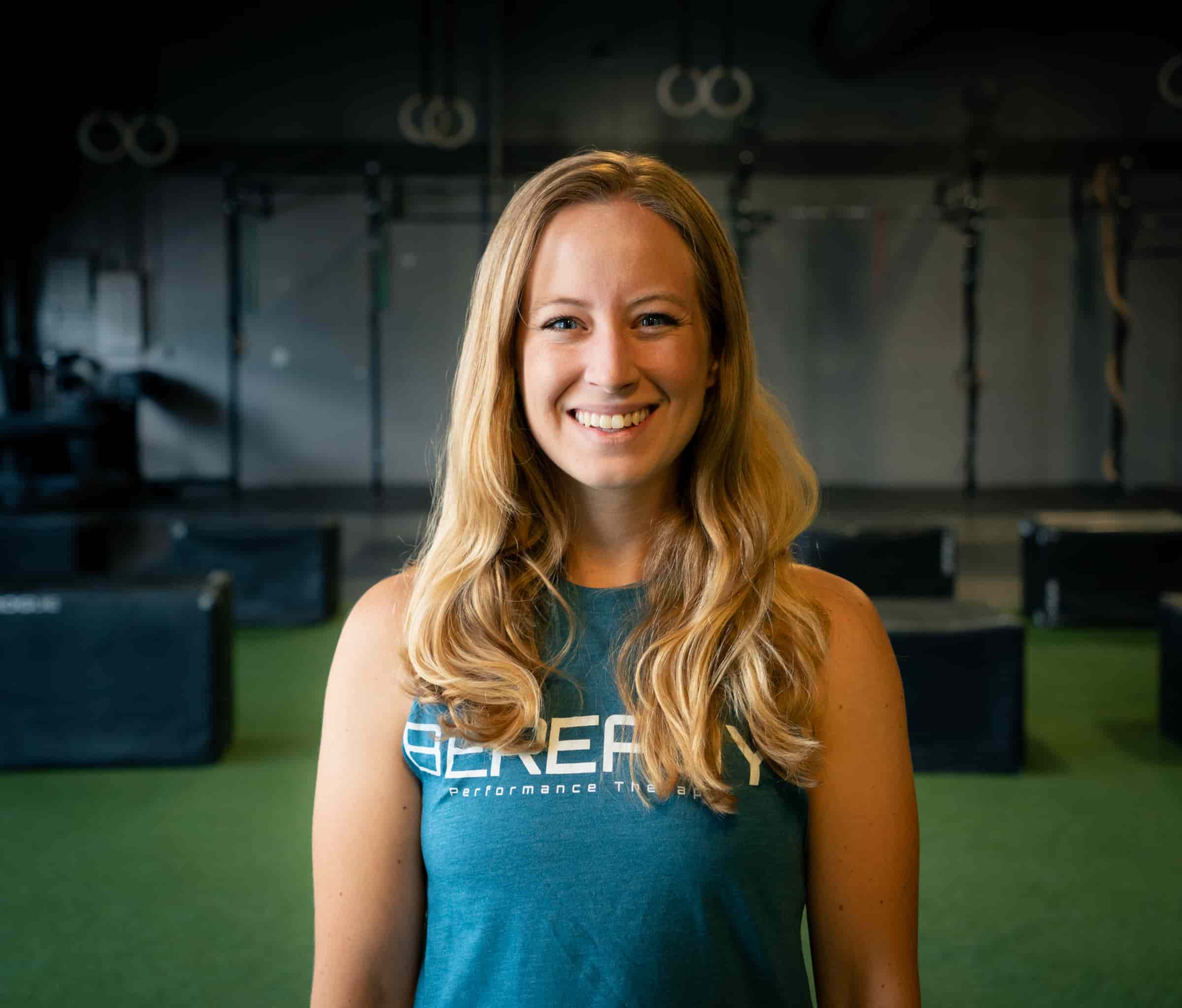A smiling woman with long blonde hair, wearing a blue tank top, stands in a gym with exercise equipment in the background.