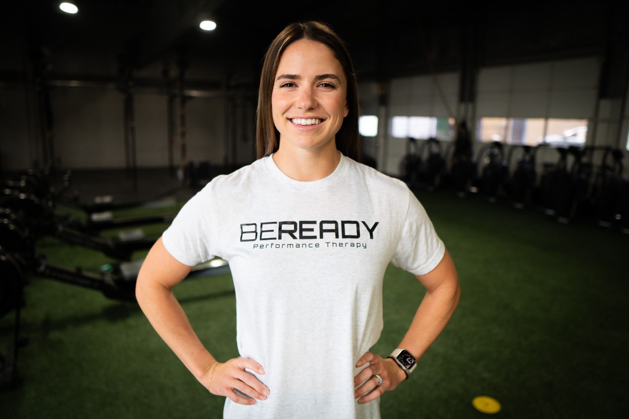 A smiling woman in a t-shirt with "beready performance therapy" printed on it, standing in a gym with exercise equipment in the background.