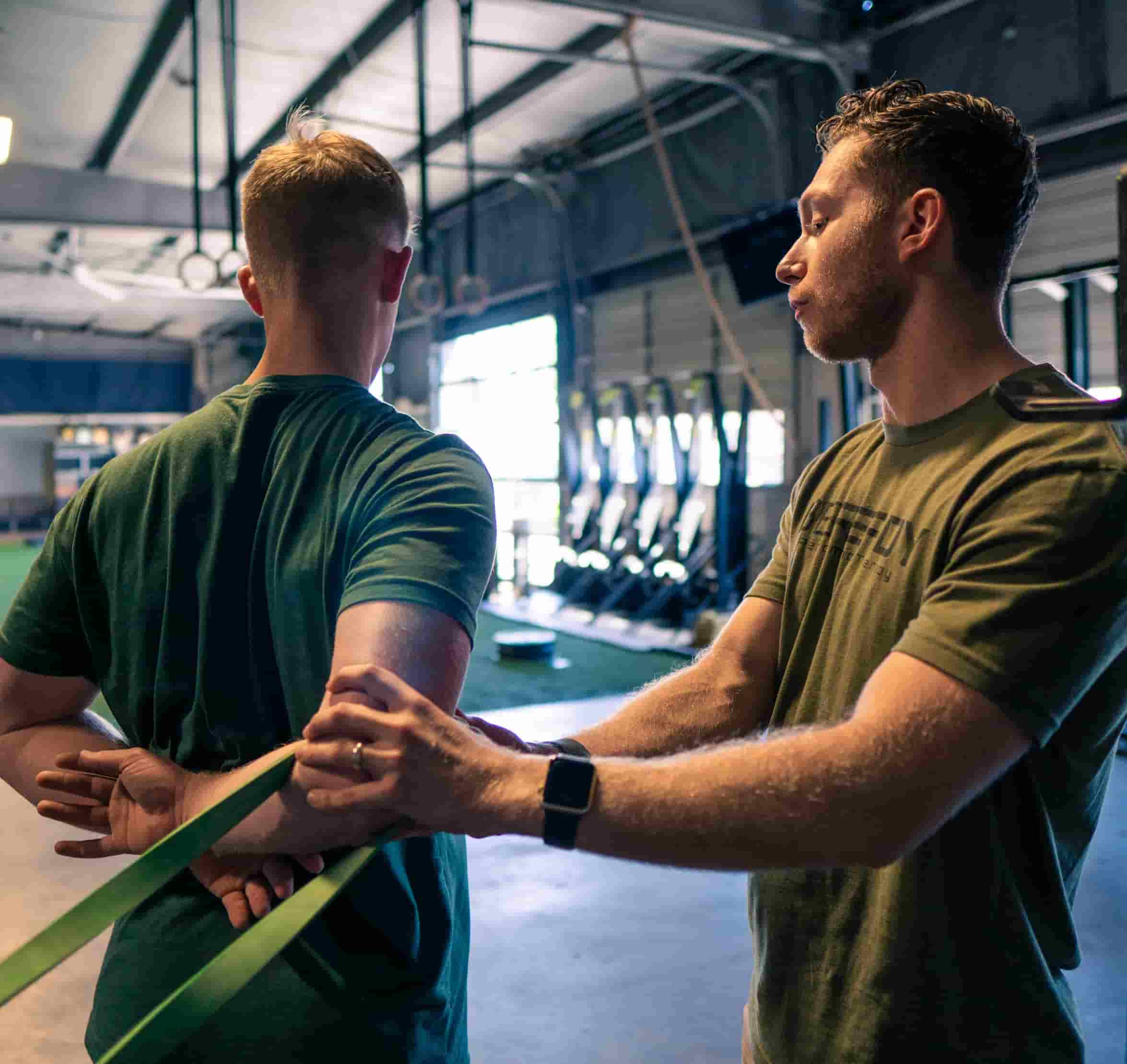 Two men in a gym, one assisting the other with stretching exercises using a resistance band, both wearing green t-shirts in a spacious fitness center.