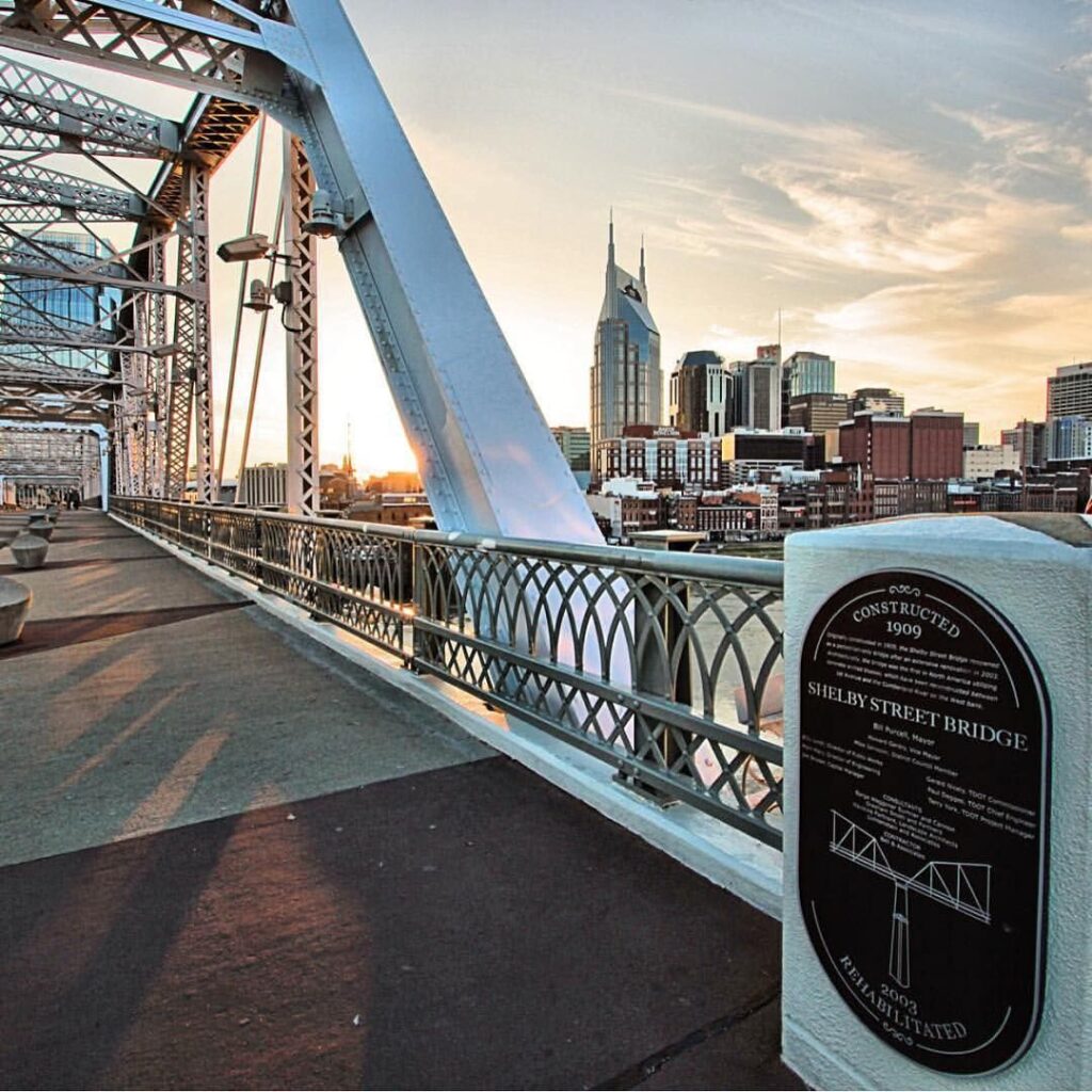 A view from the pedestrian walkway of the Shelby Street Bridge at sunset in Nashville. The background features the city skyline including the prominent AT&T building. A historical plaque is visible on the right side of the image.